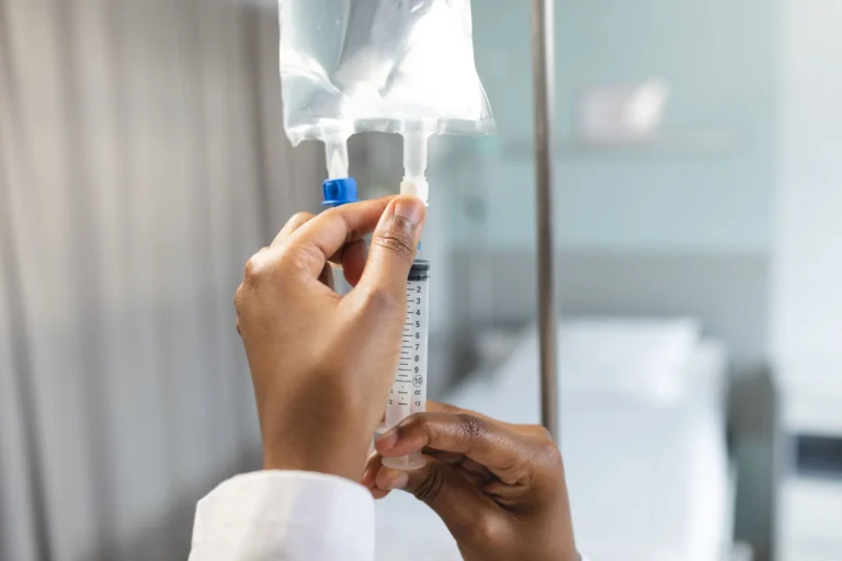 Fotografia em close-focado nas mãos de uma médica negra, vestindo jaleco branco, manuseando com precisão uma seringa acoplada a um equipo de soro em um quarto de hospital. Ao fundo, de forma desfocada, observa-se um leito hospitalar e cortinas claras, transmitindo um ambiente de cuidado clínico profissional e higienizado.
