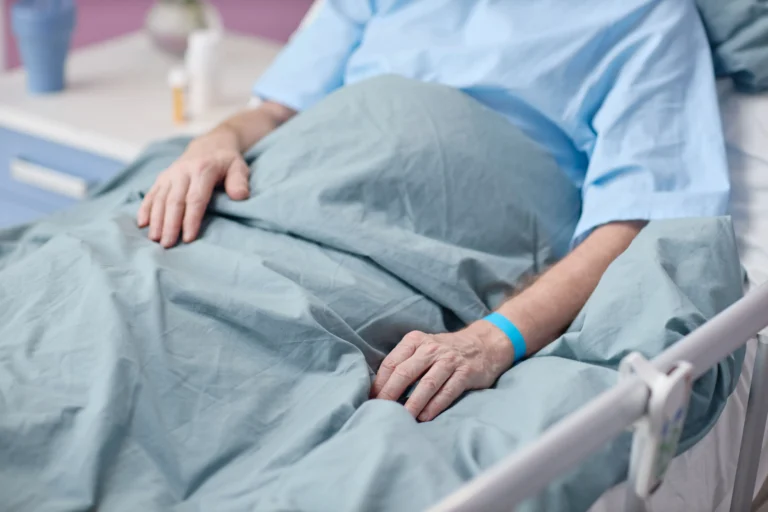 Foto de um paciente idoso deitado em uma cama de hospital, coberto por um lençol azul-acinzentado. O paciente veste uma bata azul e possui uma pulseira de identificação azul no pulso. Suas mãos estão repousadas sobre o lençol. Ao fundo, em uma mesa de cabeceira branca, veem-se alguns frascos de medicamentos. A imagem transmite um ambiente de cuidado clínico e recuperação.
