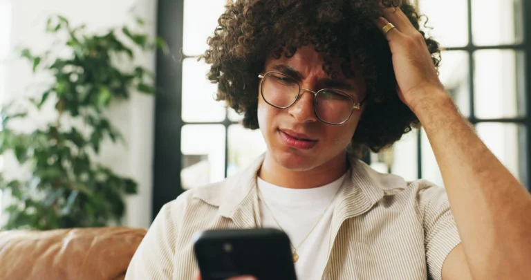 Homem jovem com expressão confusa e preocupada olha para a tela do celular enquanto coça a cabeça, ilustrando a reação de um beneficiário ao descobrir que o seu plano de saude foi cancelado de surpresa.