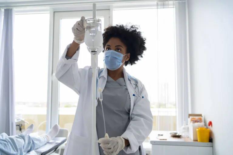 Médica negra com máscara cirúrgica, jaleco branco e estetoscópio, ajustando cuidadosamente um gotejador de soro com Kyprolis ou infusão intravenosa com Kyprolis em um ambiente hospitalar iluminado, com um paciente deitado ao fundo.
