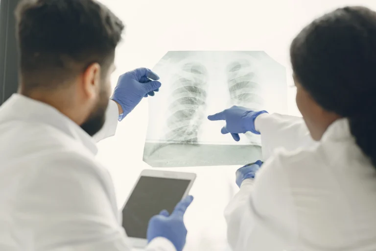 Dois médicos especialistas analisando detalhadamente um exame de radiografia do tórax de um paciente contra a luz, identificando a necessidade de intervenção respiratória e avaliando a indicação cirúrgica para desobstrução das vias aéreas com a colocação de um stent traqueal ou brônquico para reestabelecer o fluxo de ar nos pulmões.