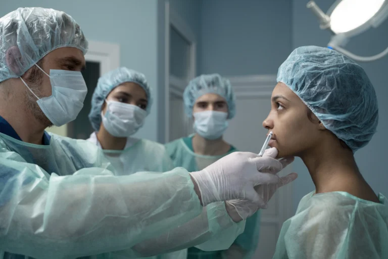 Médico cirurgião de máscara e luvas avaliando o rosto de uma paciente jovem antes de uma cirurgia plástica, acompanhado por sua equipe médica ao fundo em ambiente cirúrgico.