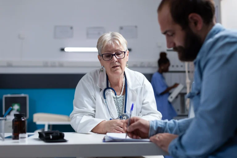 Um homem de perfil, com barba e camisa jeans azul clara, assina um documento com uma caneta azul sobre uma mesa branca de consultório médico. Do outro lado da mesa, uma médica mais velha, de óculos, cabelos curtos e grisalhos, vestindo jaleco branco e com um estetoscópio azul no pescoço, observa atentamente a assinatura dele. Ao fundo, uma enfermeira negra de uniforme azul mexe em uma pasta de arquivos, e o ambiente clínico exibe tons de azul e branco nas paredes, com alguns frascos e equipamentos médicos sobre a mesa em primeiro plano.