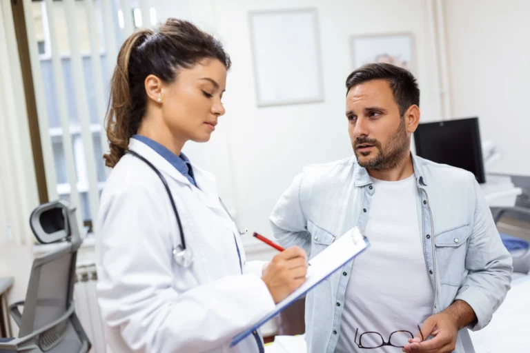 Uma médica jovem de cabelos presos e jaleco branco segura uma prancheta azul e anota informações importantes sobre a indicação de pancreatectomia enquanto ouve atentamente um paciente homem sentado em um consultório de hospital.