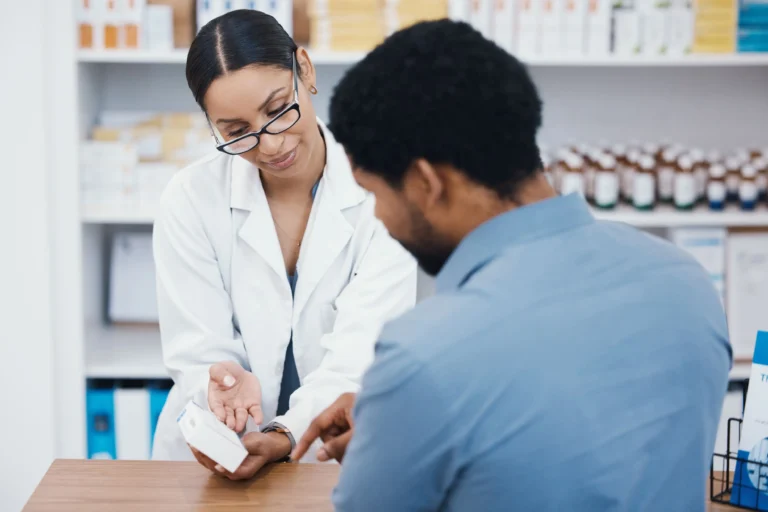 Fotografia em plano médio com foco em uma farmacêutica, mulher negra de óculos e jaleco branco, explicando como conseguir remédio pelo SUS fora da lista do RENAME ao analisar uma caixa de medicamento com um cliente, homem negro visto de costas. Ao fundo, prateleiras brancas com diversos produtos de saúde estão levemente desfocadas.