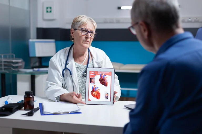 Médica cardiologista de jaleco explica diagnóstico de pericardiectomia mostrando a ilustração anatômica do sistema cardiovascular e a estrutura do coração em um tablet para o paciente idoso no consultório.