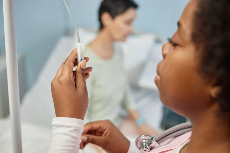 Uma profissional de saúde negra ajusta o gotejamento de uma medicação intravenosa, como o Tarlatamabe, em um ambiente hospitalar. Em primeiro plano, as mãos da profissional manipulam o equipo de infusão, enquanto ao fundo, de forma desfocada, uma paciente aguarda sentada na cama do hospital para iniciar o seu tratamento oncológico. A imagem reflete o cuidado técnico e humano necessário durante a administração de terapias biológicas.