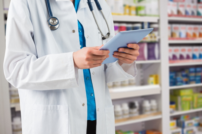 Profissional de saúde em uma farmácia segurando um tablet digital para verificar a disponibilidade e os direitos de liberação do medicamento Rinvoq de alto custo nas prateleiras ao fundo.