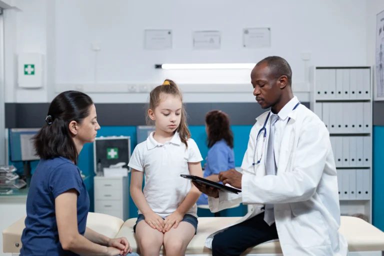 Fotografia de uma consulta médica realista em um consultório clínico hospitalar claro e bem iluminado. À direita, um médico pediatra negro, vestindo jaleco branco e estetoscópio azul no pescoço, está sentado segurando um tablet eletrônico preto enquanto digita e explica os detalhes sobre a epifisiodese. No centro, uma menina branca de cabelo castanho amarrado em um rabo de cavalo alto, vestindo uma camiseta polo branca e shorts jeans, está sentada na maca de exames estofada olhando atentamente para o tablet. À esquerda, a mãe da criança, uma mulher branca de cabelo escuro amarrado, vestindo camiseta azul-escura, observa a explicação do médico com uma expressão concentrada. Ao fundo, há elementos típicos de um consultório, como armários organizadores brancos, um monitor de computador e outra profissional de saúde com uniforme azul trabalhando distanciada.